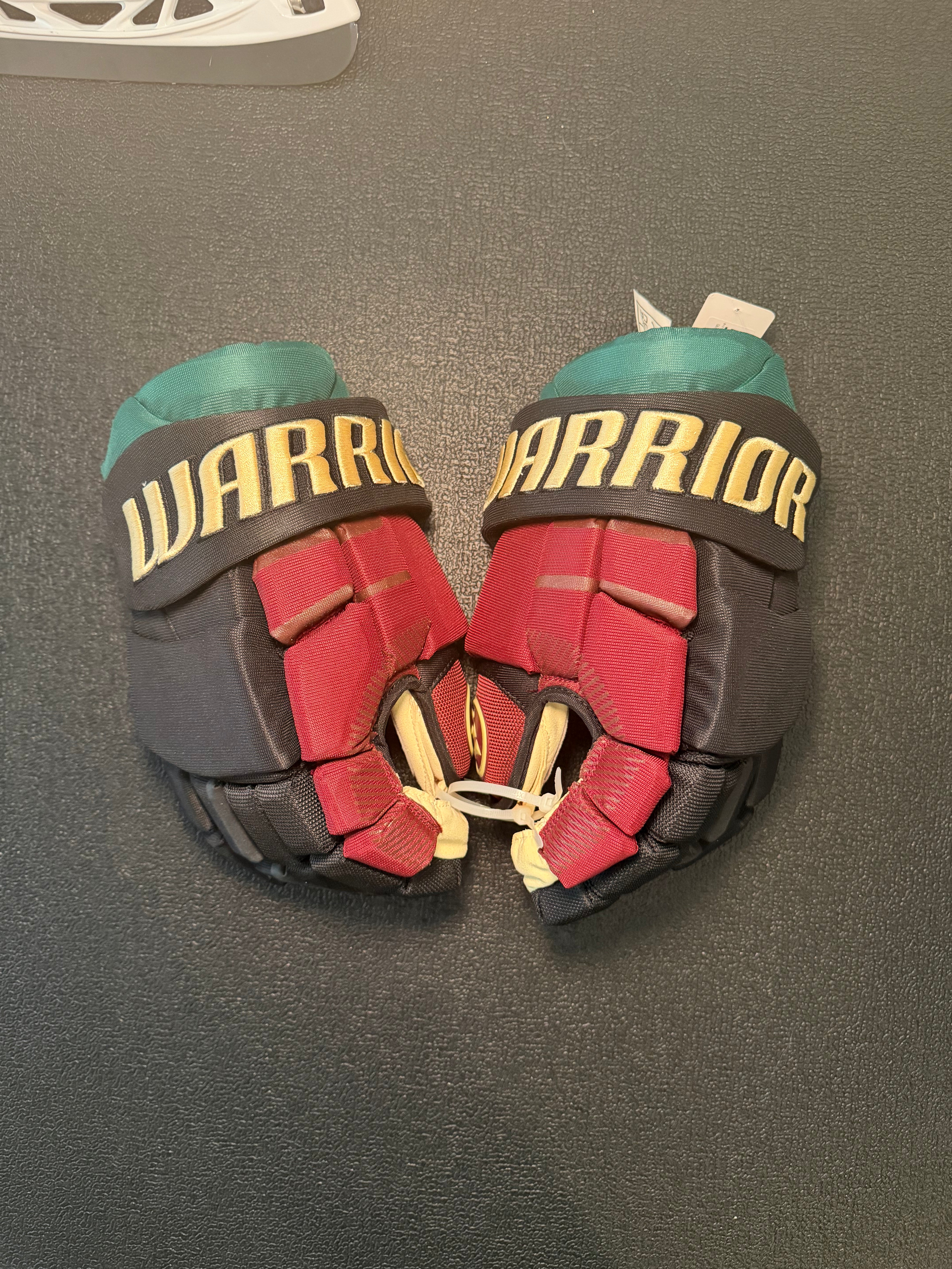 A pair of Warrior hockey gloves in red, black, and teal, laid flat on a dark gray textured surface. The gloves feature the 'WARRIOR' brand name in gold lettering across the black wrist straps.